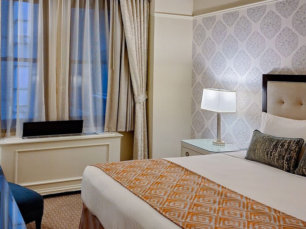 Close-up of king bed with patterned throw and beside window with curtains and desk lamp in Premier Room at Warwick New York