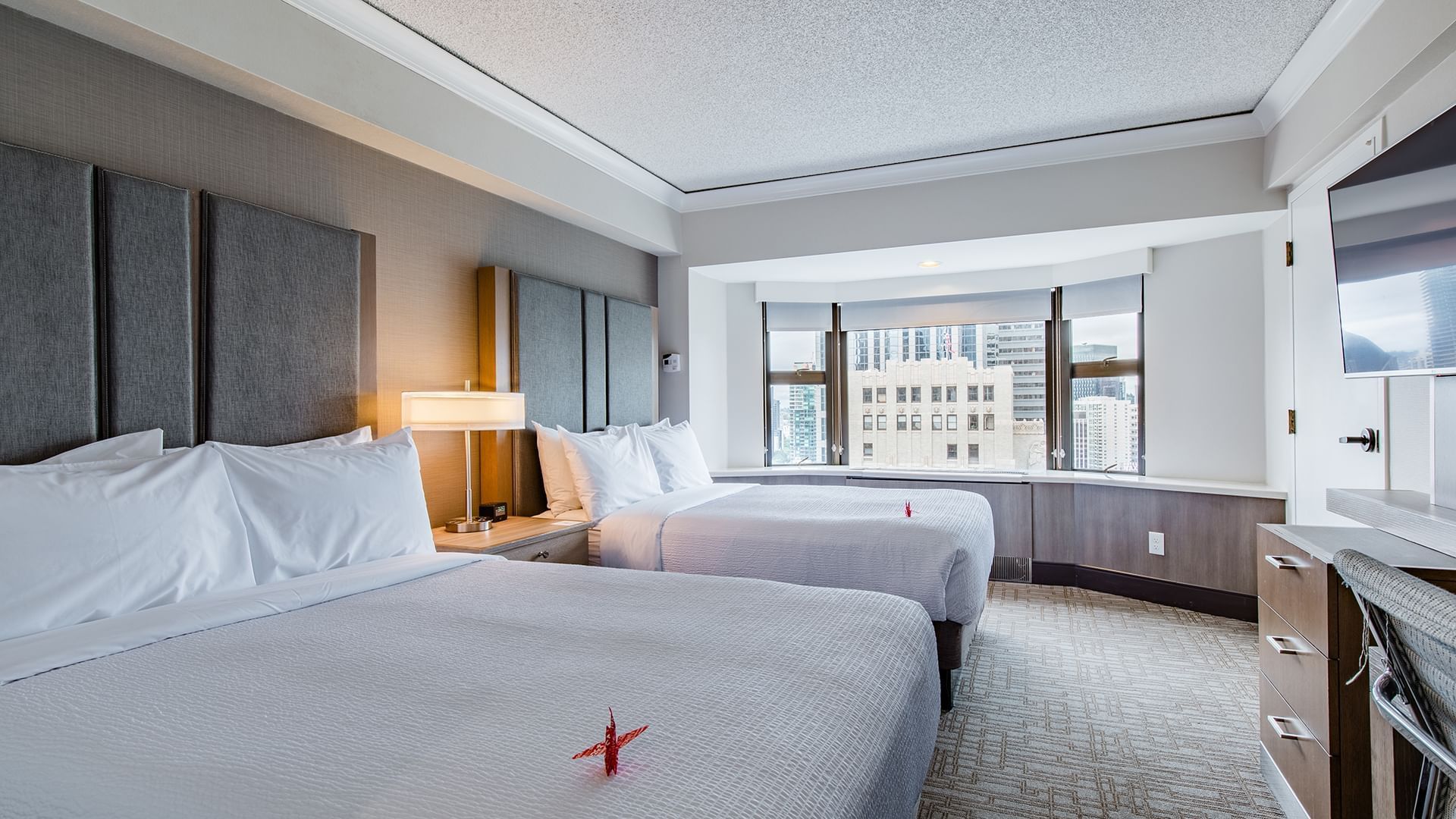 A hotel room with two beds, a window with a city view, and a desk.