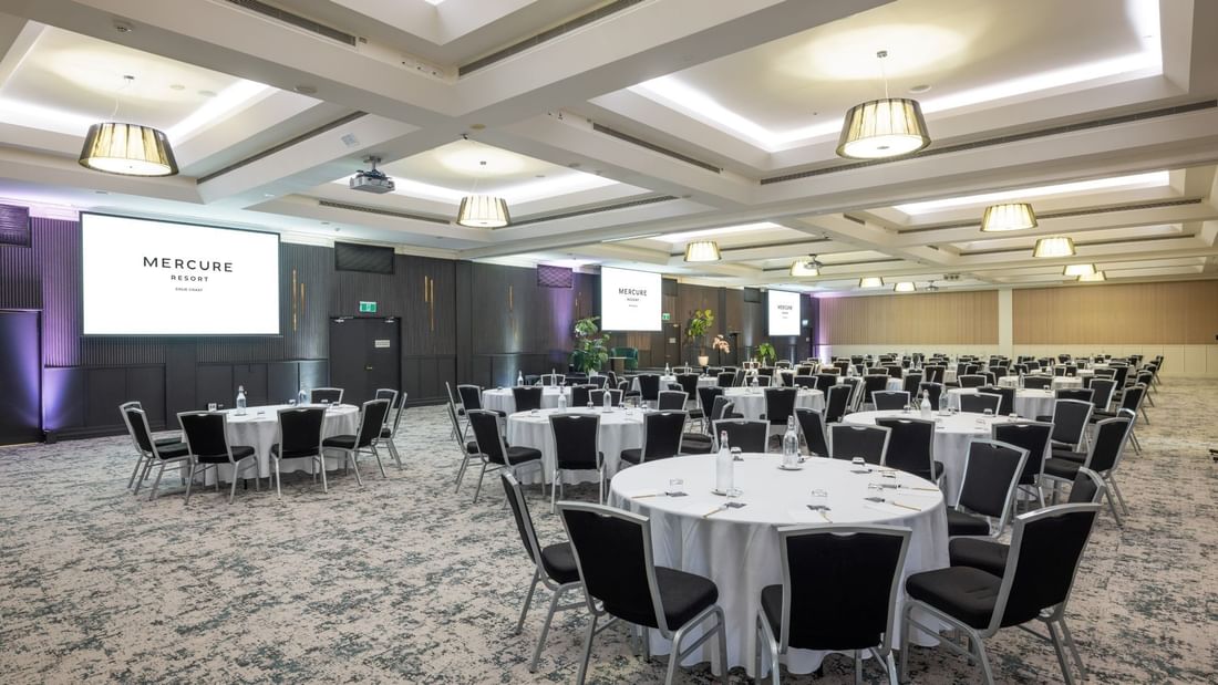 A large conference room with round tables, black chairs, and white tablecloths, with a 