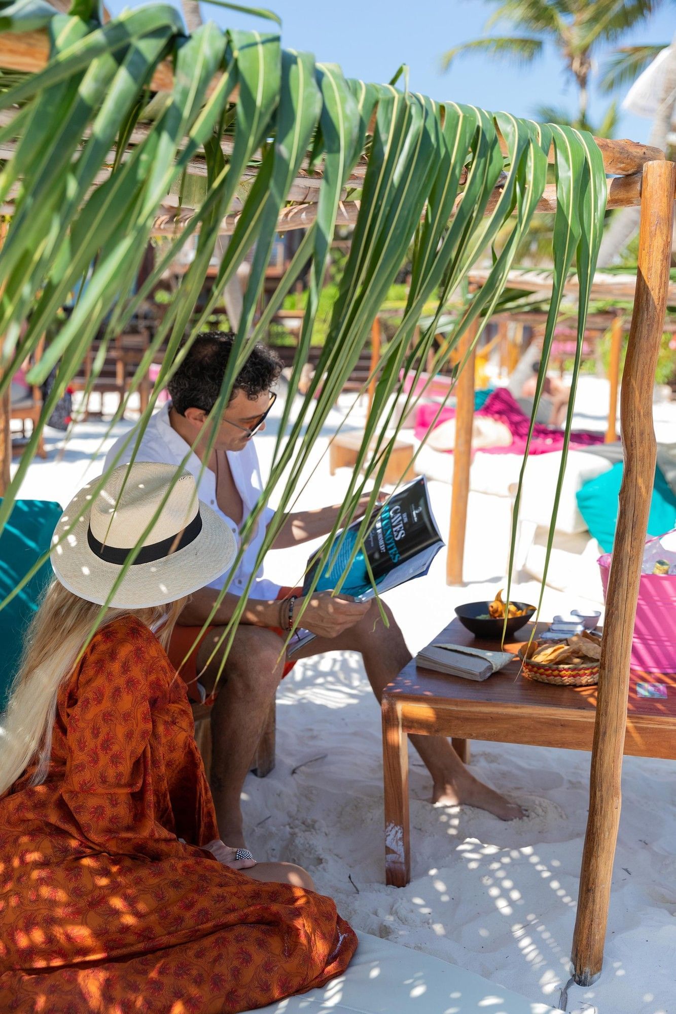 A couple reads under a palm frond cabana on the white sand beach at the vibrant La Zebra Hotel