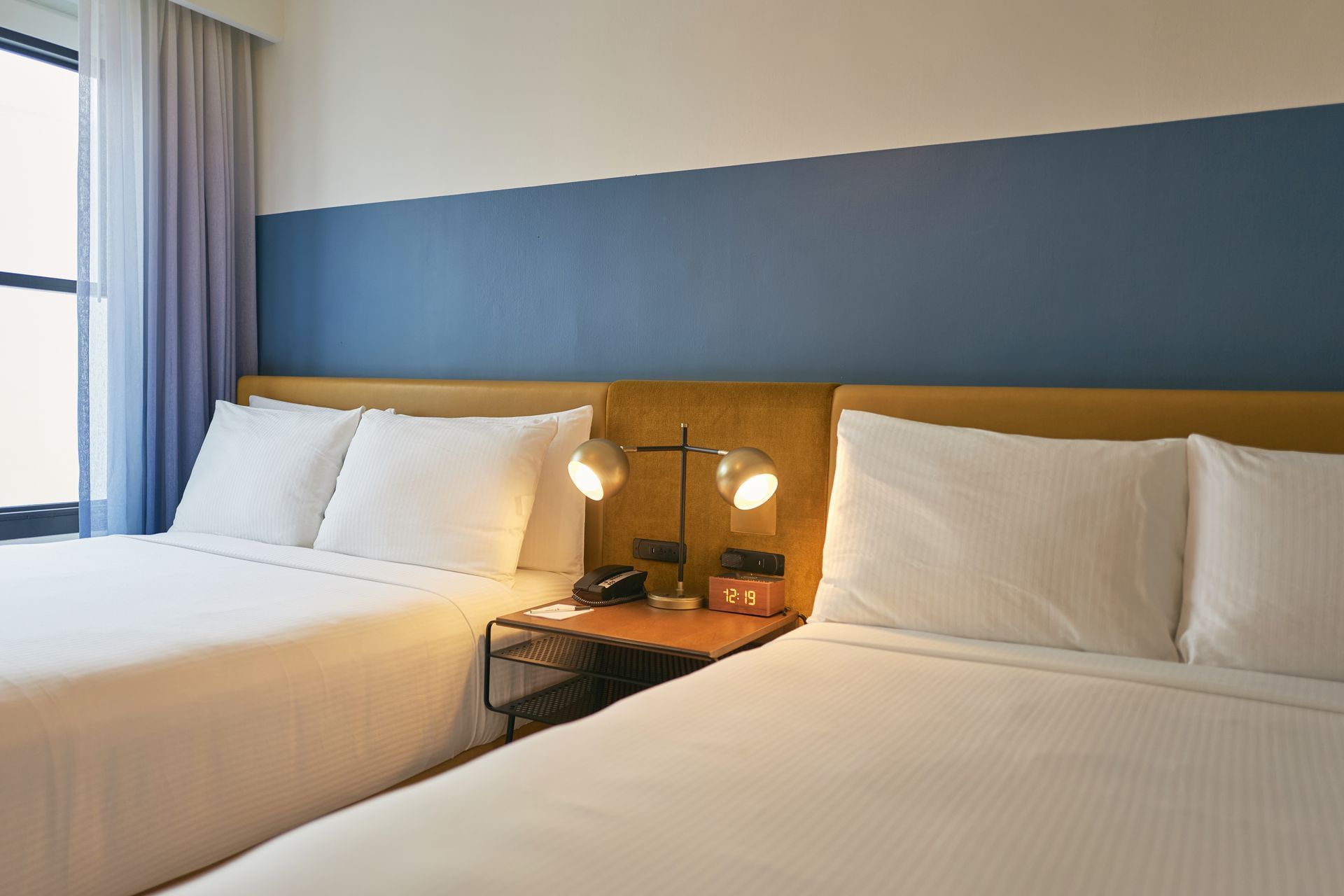 A hotel room with two double beds, a nightstand with a lamp and alarm clock, and a window with curtains.