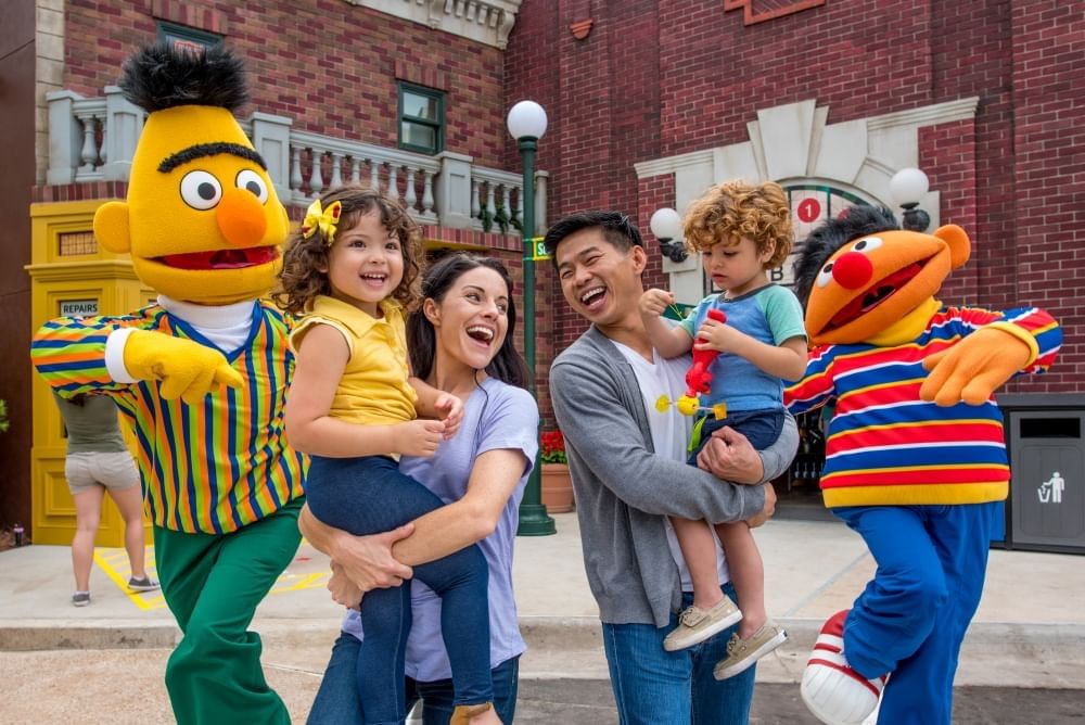 A man and woman, each holding a small child, smile at each other as they walk between Bert and Ernie from Sesame Street.