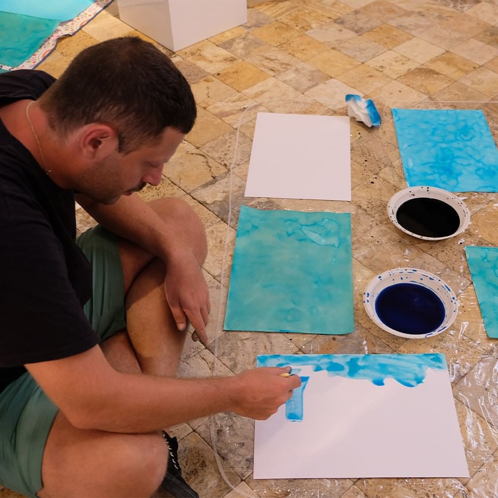Man painting blue watercolor on a white paper with other papers and paint bowls around him.