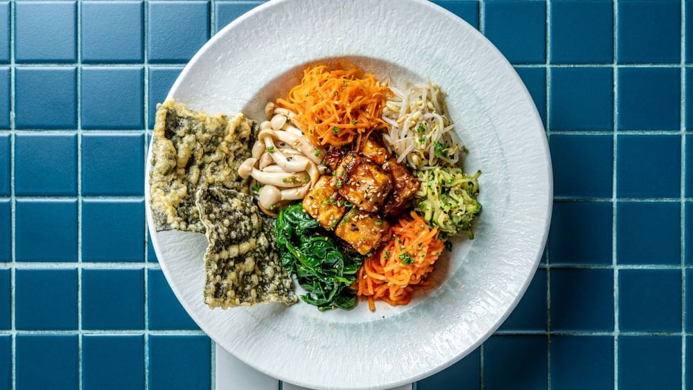 A white bowl filled with glazed tofu, spiralized carrots, greens, rice, and seaweed on blue tile for Bledisloe Cup Dining
