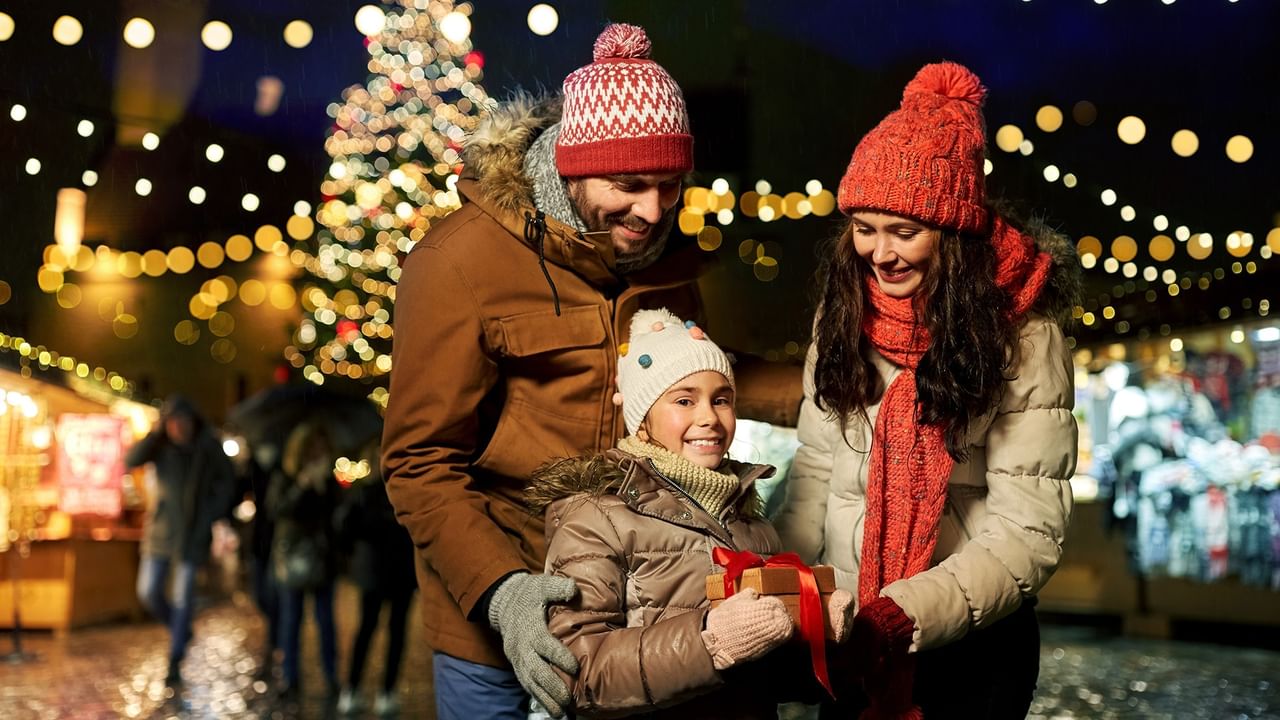 Family of three with gifts at a Christmas market, smiling under festive lights.