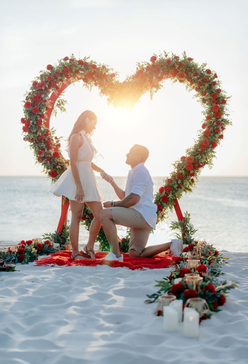 Man kneeling to propose to woman on beach under heart-shaped floral arch at sunset.