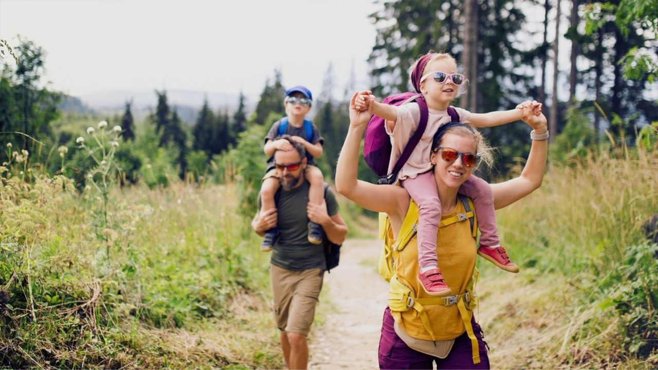 Two adults hiking with their children on a dirt path in a forest on a sunny day.