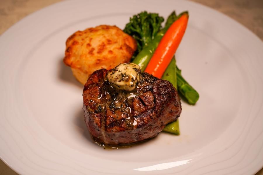 A steak topped with herb butter sits on a white plate beside a potato side and fresh colorful vegetables.