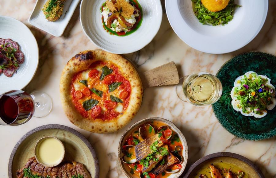 Overhead view of gourmet dishes including margherita pizza, seafood, and plated entrees on a marble table in May Fair Kitchen