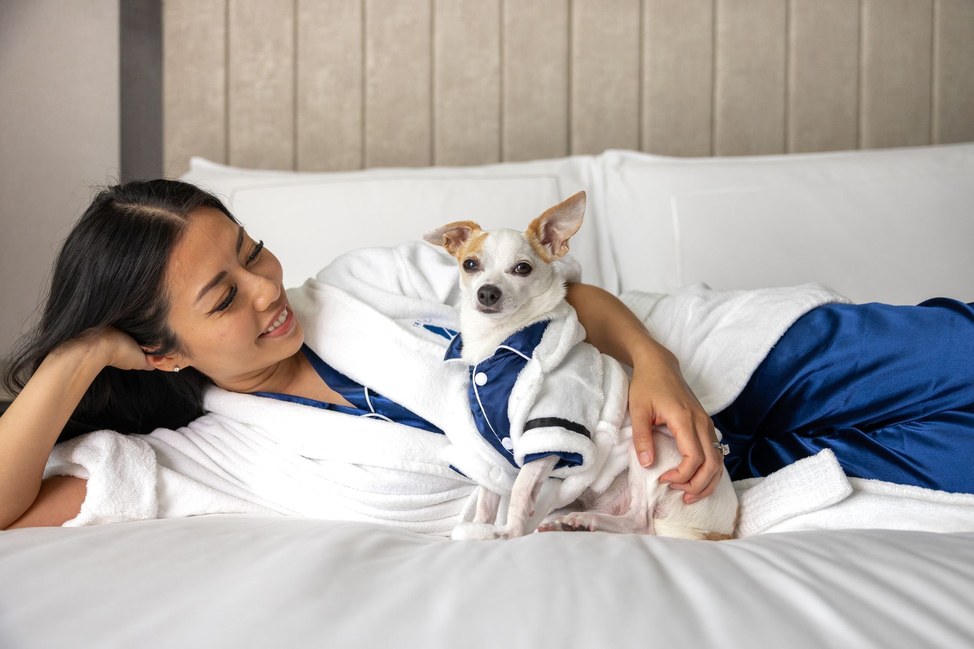 Smiling woman in white bathrobe lying on bed next to small dog wearing bathrobe and pajamas at Hotel X Toronto