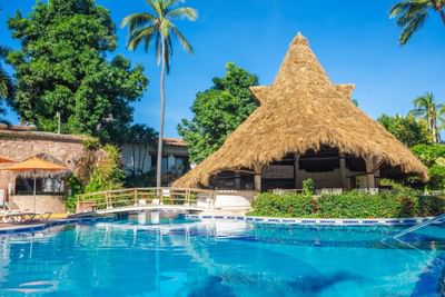 Pool with blue water, wooden bridge, thatched roof bar, and tropical trees under a clear blue sky.
