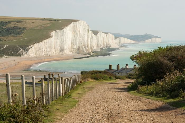 Dirt path along a fence overlooking white cliffs, a beach, and a house by the sea.