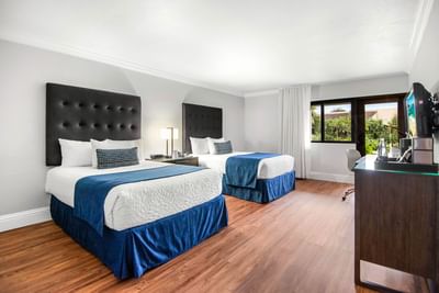 Two beds with blue throws beside a window overlooking trees and work desk in the Accessible Deluxe room at Miami Lakes Hotel