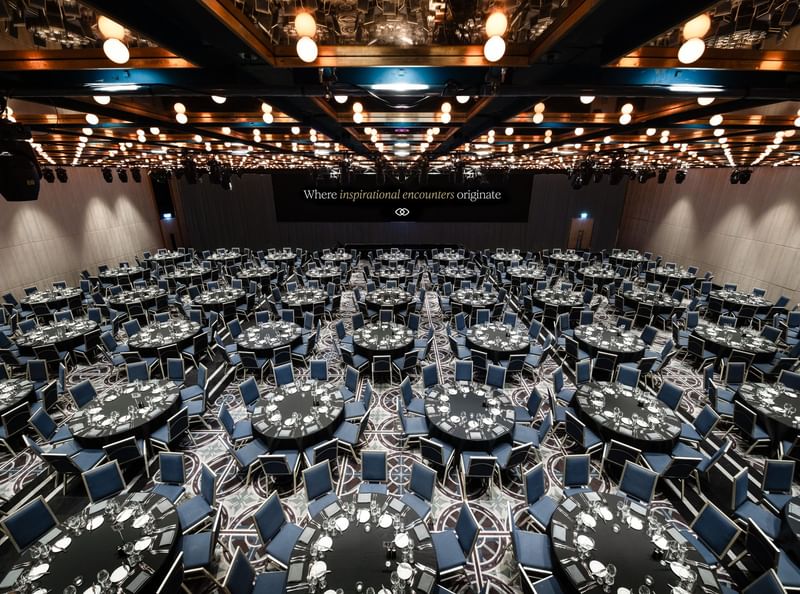 A large banquet hall with round tables and chairs set up, a stage with a large screen, and decorative lighting.