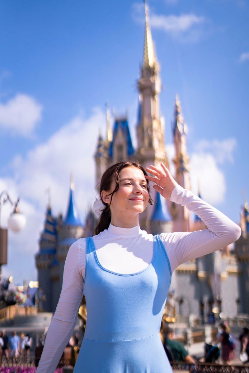 A woman with brown hair dressed up similar to Belle from Beauty and the Beast, wearing a white long-sleeve shirt and blue dress over the top. She's standing in front of the castle at Walt Disney World in Orlando.