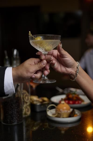 A man's hand and a woman's hand hold a martini glass in front of a dim table set with elegant out-of-focus dishes. 