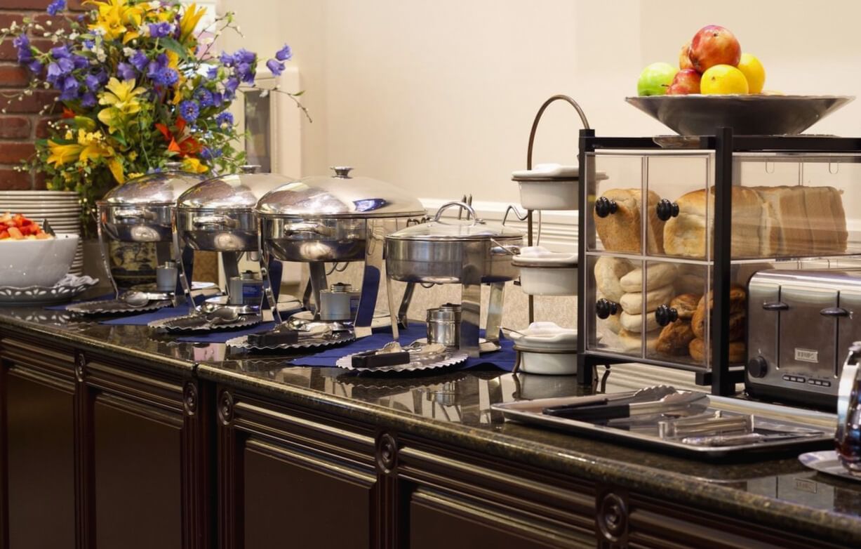 A buffet spread with chafing dishes, fruit, and bread on a marble countertop.