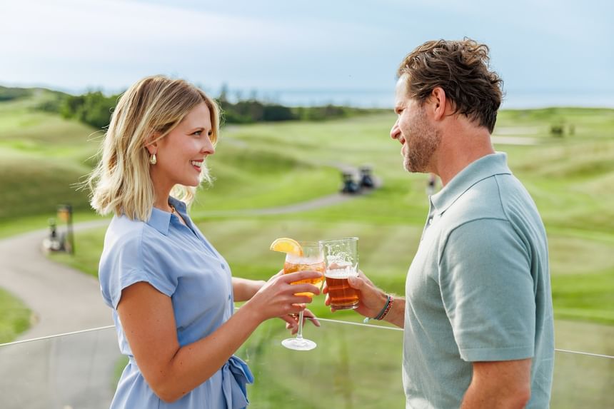 Couple toasting with drinks on a balcony overlooking the green golf course at Rodd Crowbush Golf & Beach Resort