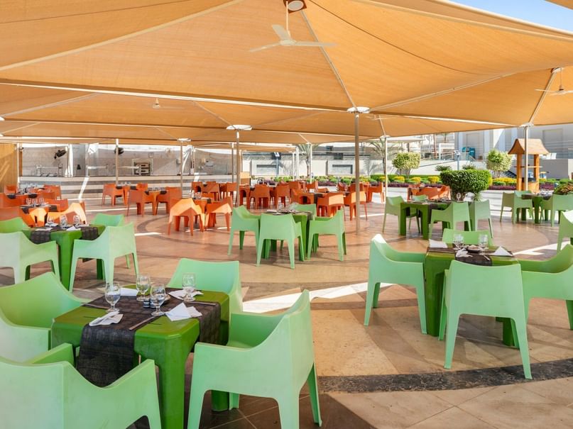 An outdoor dining area with green and orange chairs, tables, and large tan canopies.