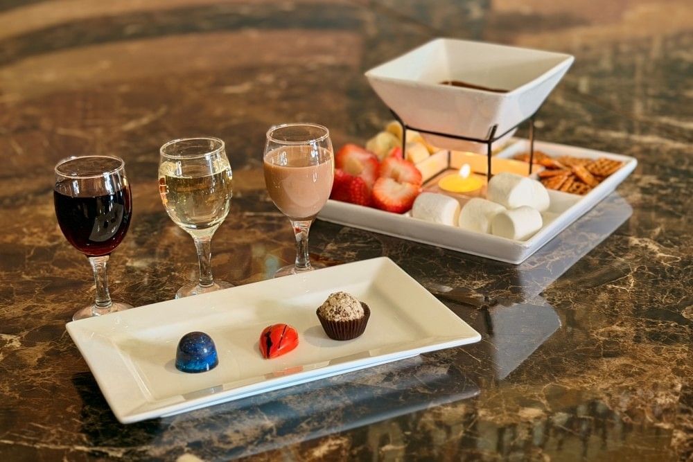 A plate with three chocolates sits on a table beside three glasses of wine and a chocolate fondue dish with marshmallows, strawberries, and other goodies. 