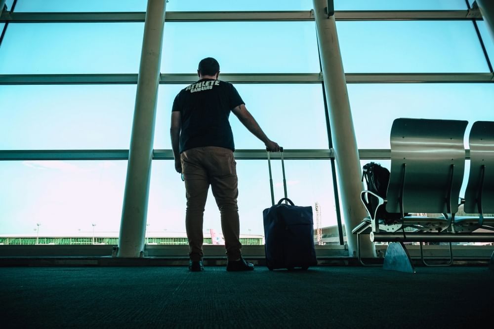 A man in a tshirt with his back to the camera and his hand on a suitcase staring out at large airport windows. Check multiple airports for cheap flights to Orlando.