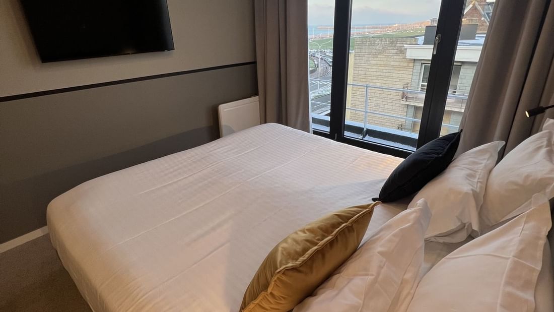 A hotel room with a large bed, TV, and windows with curtains showing a city view.