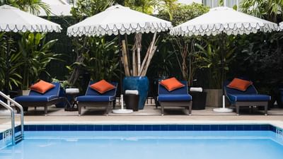 Blue poolside sun loungers with orange pillows and white umbrellas under palm trees at Riviera Hotel South Beach
