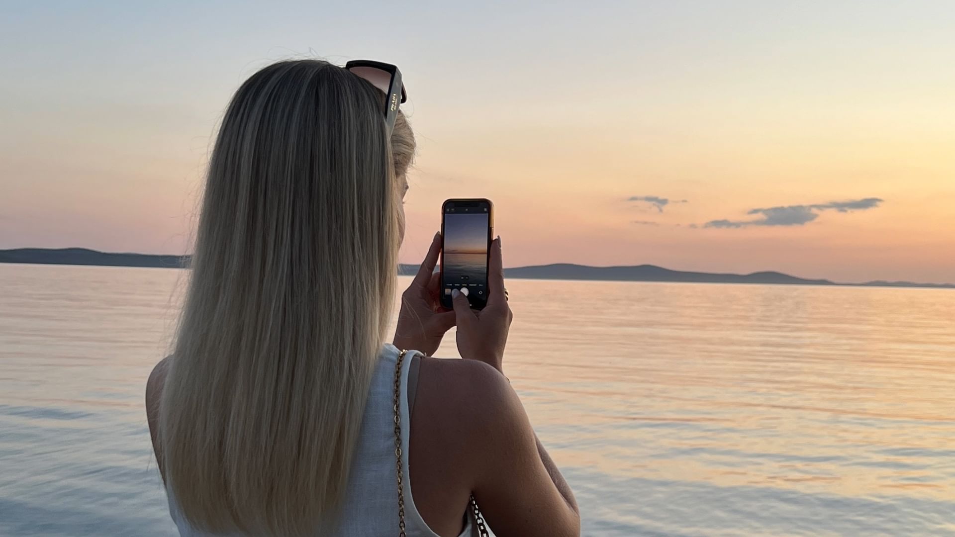 Blondine mit Sonnenbrille auf dem Kopf macht ein Foto vom Sonnenuntergang über dem Meer mit ihrem Handy.