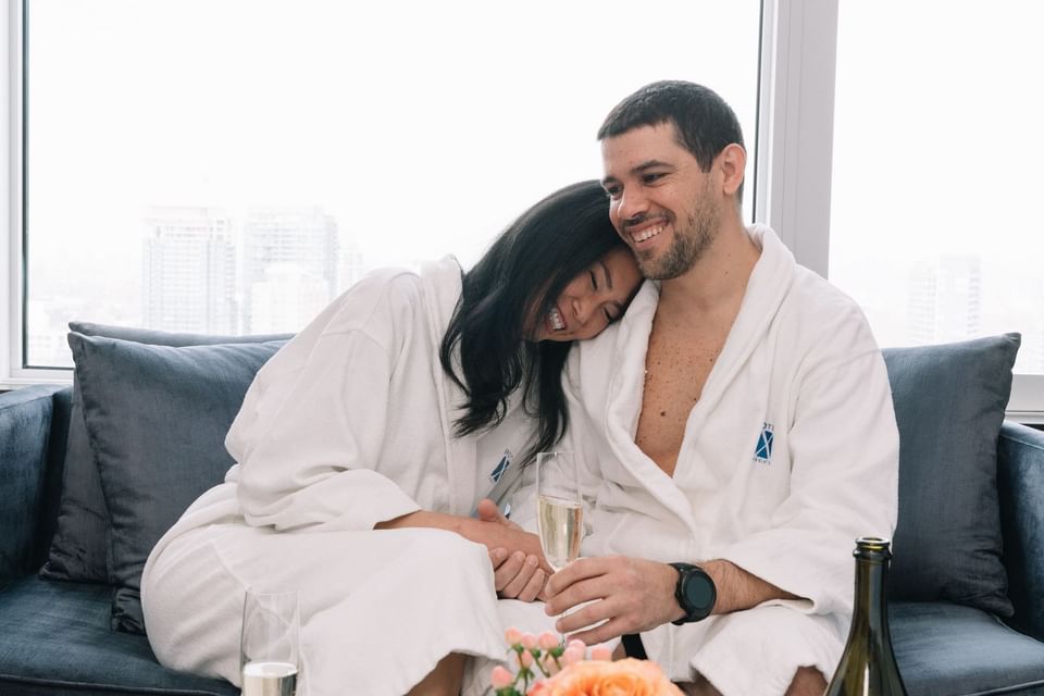 Smiling couple in white bathrobes sitting on blue sofa with champagne glasses in room at Hotel X Toronto