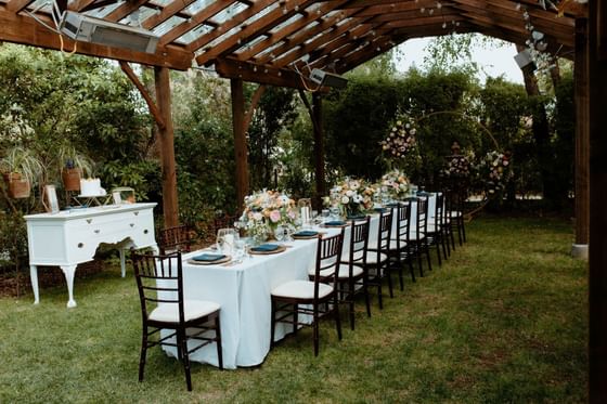Event reception long table set with white linen and wooden chairs, under a timber pergola canopy at Cambria Pines Lodge