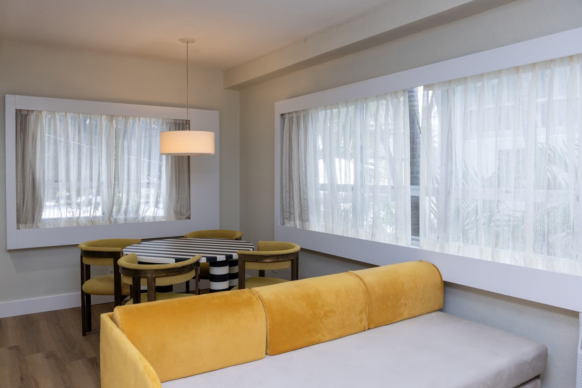A furnished room with a yellow sofa, dining table, and chairs in front of large windows.