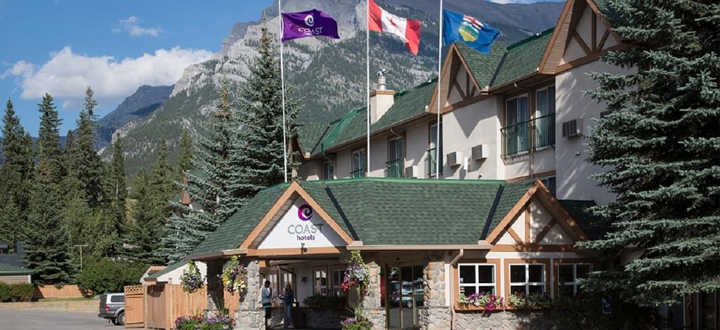 Front entrance of Coast Canmore Hotel, with Canadian and hotel flags flying and forested mountains behind.