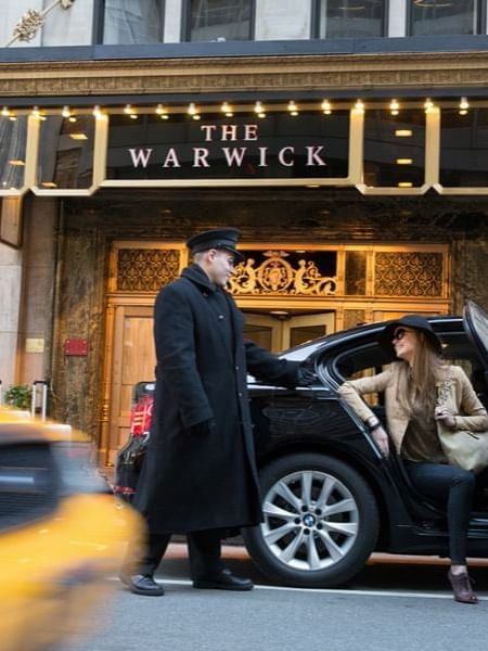 Hotel doorman holds a black sedan door open for a female guest under a lit canopy at Warwick New York