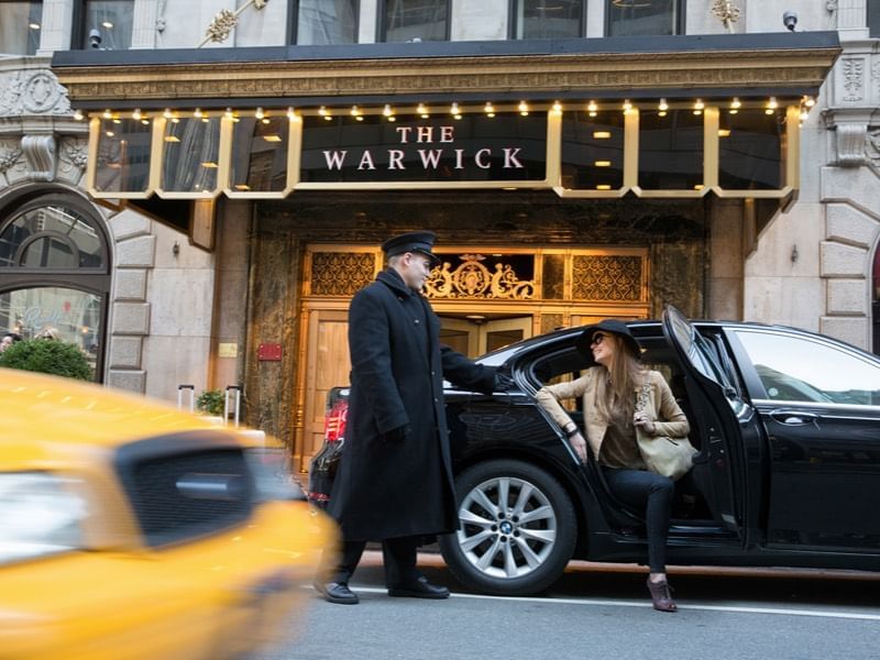 Hotel doorman holds a black sedan door open for a female guest under a lit canopy at Warwick New York