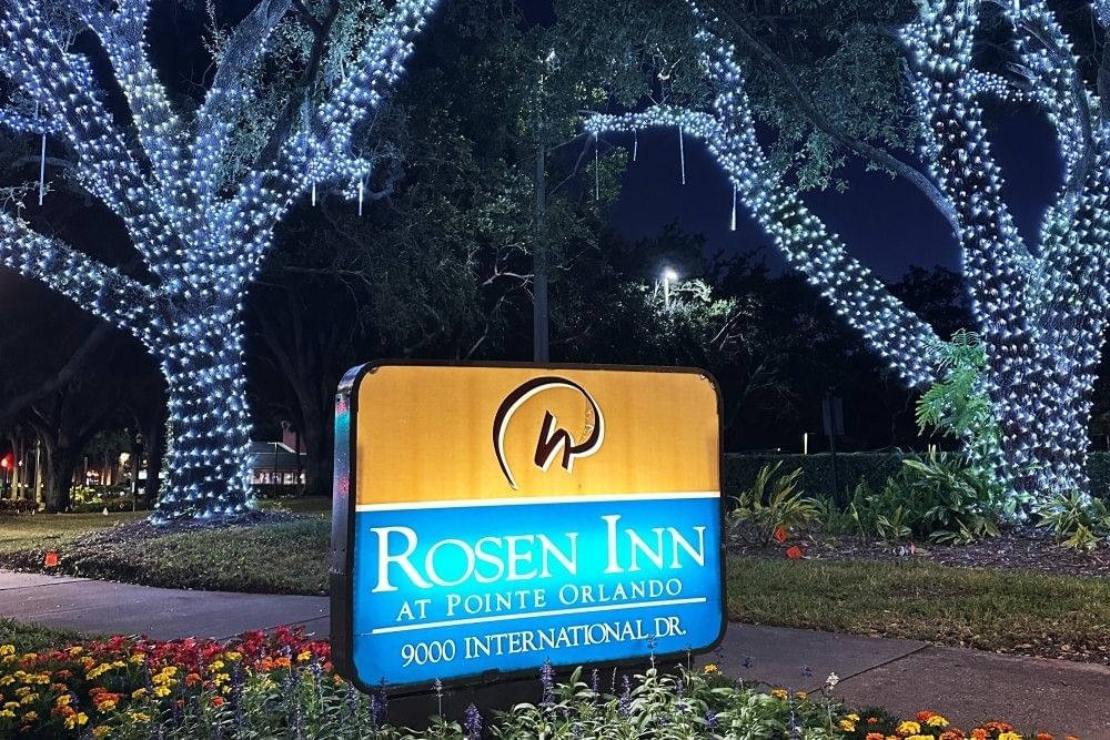 A sign for Rosen Inn at Pointe Orlando at night between two beautiful trees wrapped in white Christmas lights.