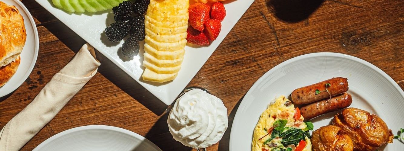 Plates of fruit, omelets, and sausages on a wooden table in In-Room Dining at Warwick Denver