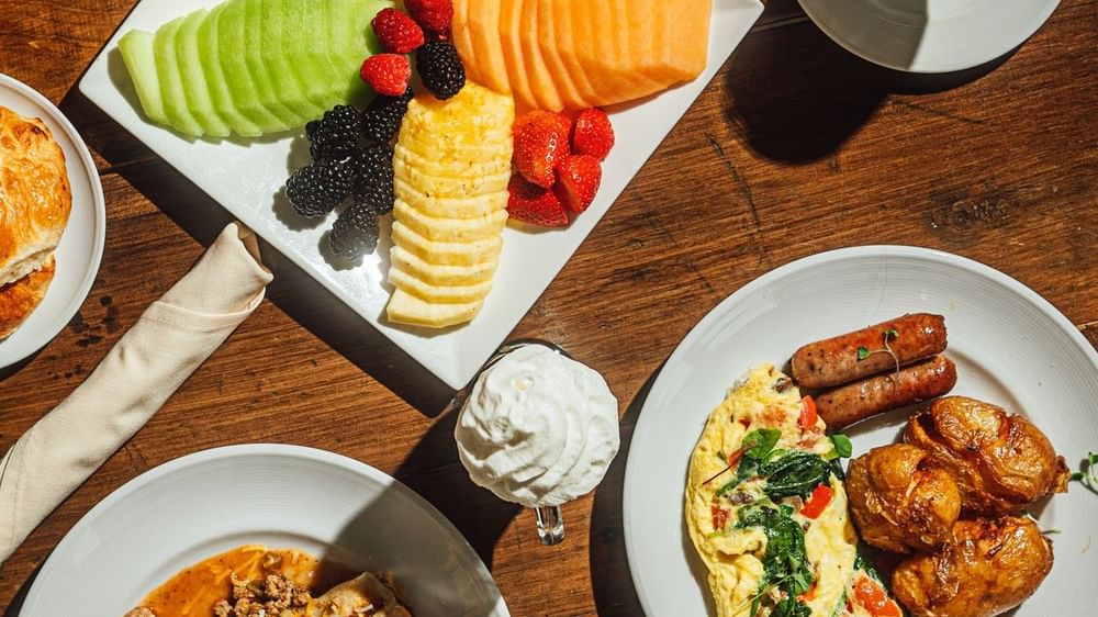 Plates of fruit, omelets, and sausages on a wooden table in In-Room Dining at Warwick Denver