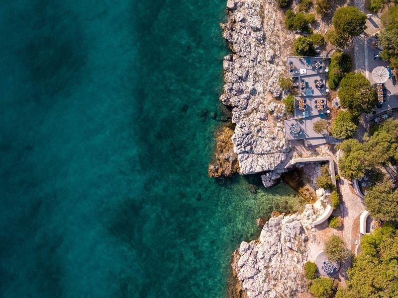 Vista aerea di una costa rocciosa con una terrazza e un percorso lastricato tra gli alberi.