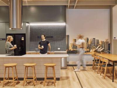 Interior of a modern kitchen with three people, wooden furniture, and a cityscape mural.