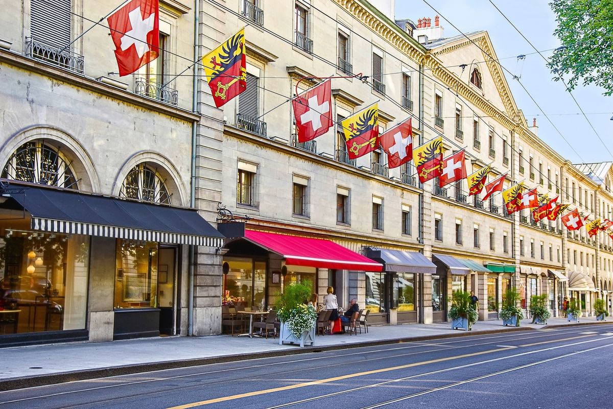 Swiss flags hanging by a city building under a blue sky near outdoor cafes near Warwick Geneva