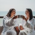 Two women in white robes with hotel logo, holding mugs, laughing on a balcony overlooking the ocean.