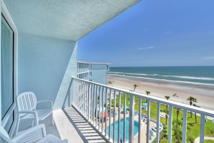 A balcony with white chairs and a railing overlooking the ocean, pool, and beach.
