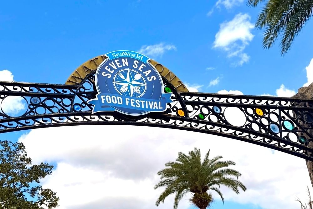 A metal arch with a sign for SeaWorld Orlando's Seven Seas Food Festival against a blue and cloudy sky.