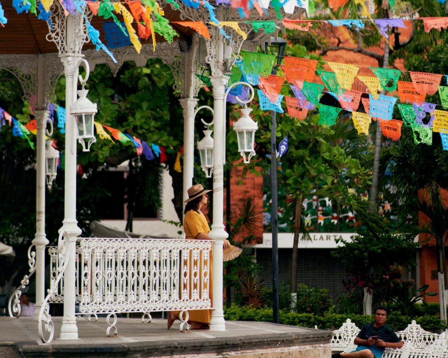 Mujer observando el quiosco tradicional decorado con papel picado en la plaza del centro de Puerto Vallarta, rodeado de vegetación y ambiente cultural mexicano.