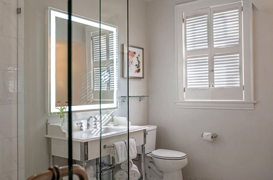 White vanity by a toilet under a lighted mirror surrounding a marble bathroom in Deluxe Suite at Warwick Melrose Dallas