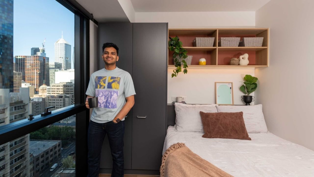 A resident stands in a bedroom with a bed, cityscape view, and organized shelving.