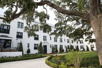 White building with black windows at The White House Hotel under large oak trees and a green lawn on a sunny day