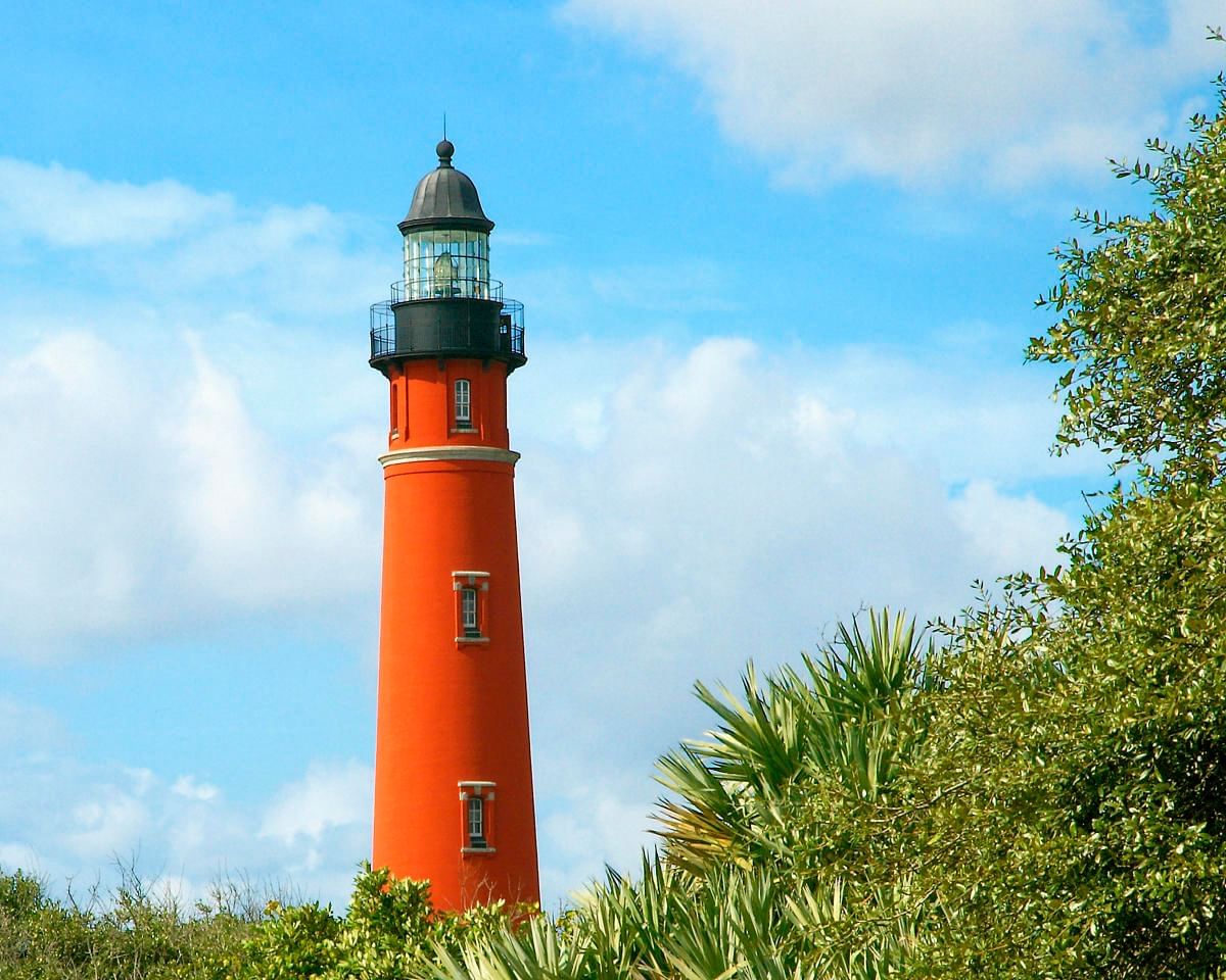 A bright orange lighthouse with a black roof and glass enclosure at the top, surrounded by green trees and a blue sky with white clouds.