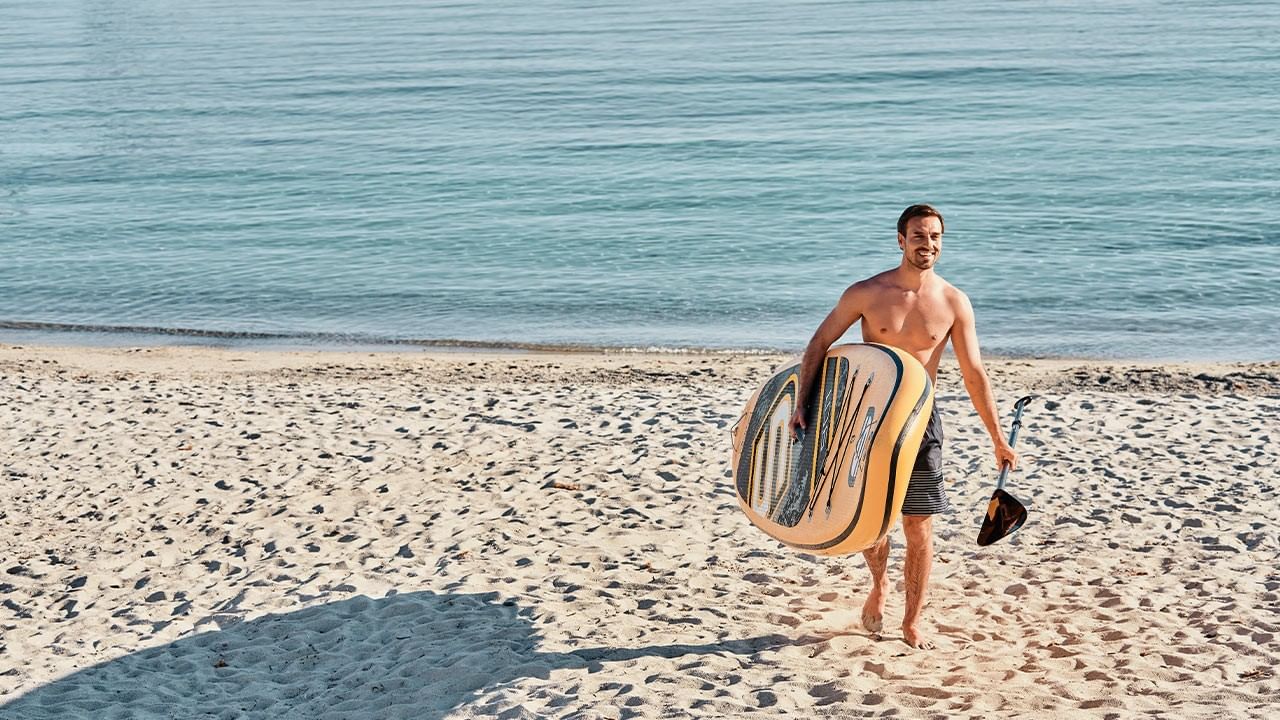 Man holding paddle and inflatable float, walking towards ocean on sandy beach.