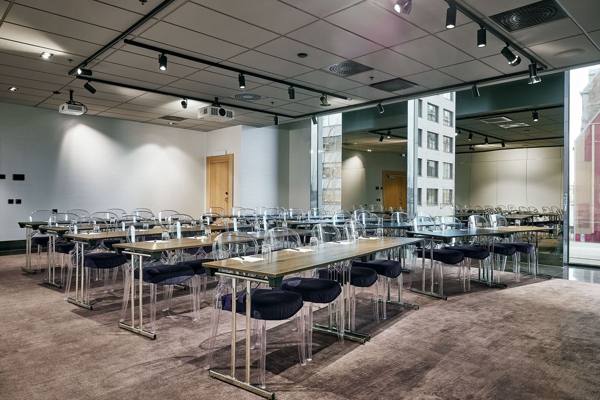 Konferensrum med två projektorer, bord och stolar, och stora fönster med utsikt över ett högt byggnad.